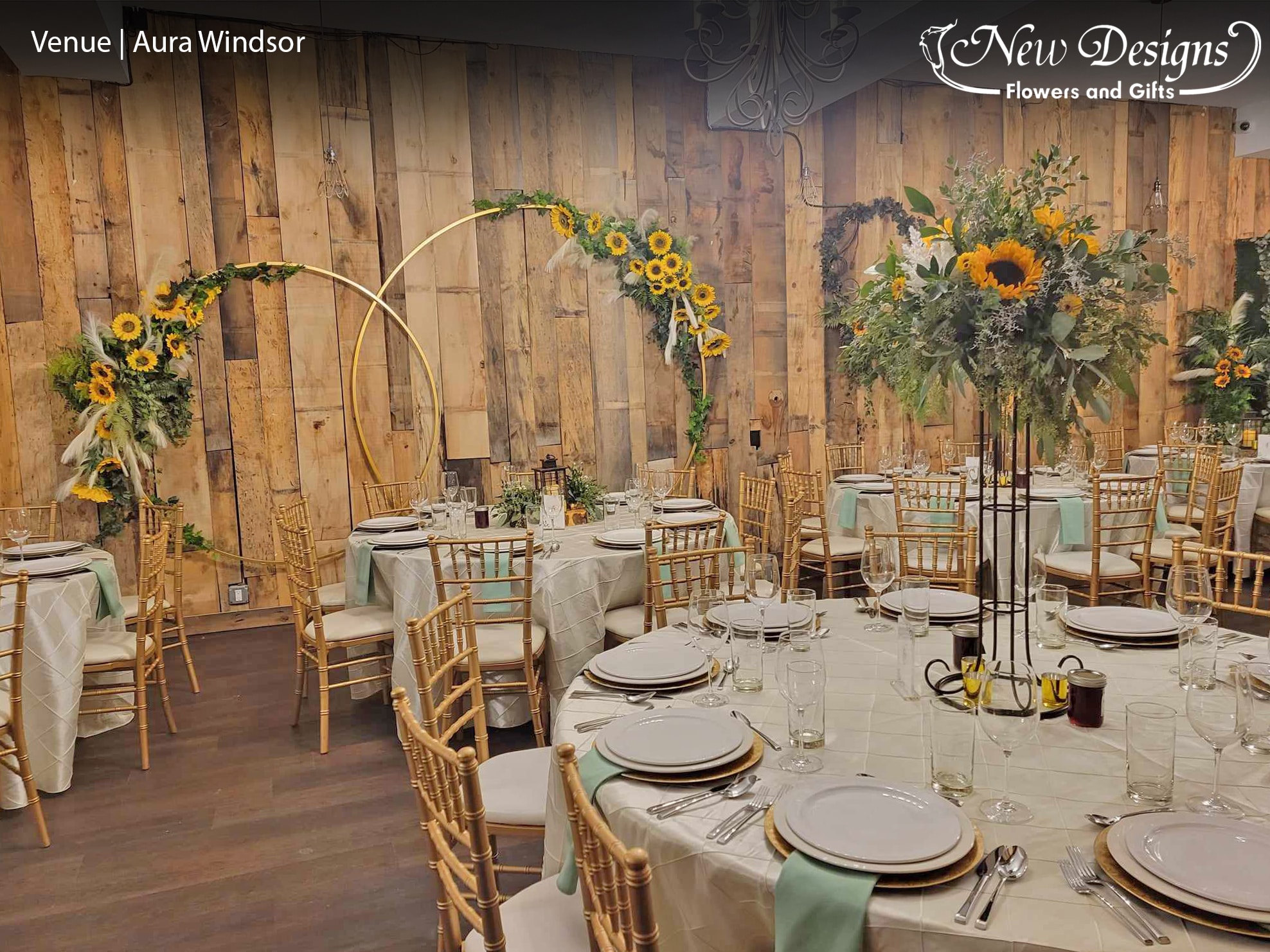 A gorgeous rustic bar venue filled with Greenery and Sunflowers. Centerpieces sitting atop tall 4 posted Iron frames. Gold Hoops wrapped with greenery and sunflowers Scatter at various heights along a barnwood wall,